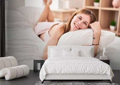 Young blonde woman hugging pillow lying on bed smiling at bedroom Wall mural