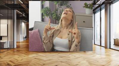 Young blonde woman in an indoor apartment living room, pointing upwards with both hands while sitting on a couch, surrounded by decor and houseplants, exuding a joyful expression. Wall mural