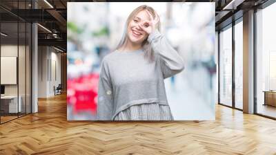 Young blonde woman over isolated background doing ok gesture with hand smiling, eye looking through fingers with happy face. Wall mural