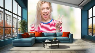 Young blonde woman over isolated background smiling with happy face winking at the camera doing victory sign. Number two. Wall mural