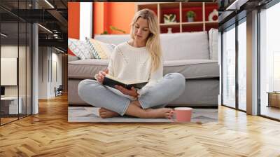 Young blonde woman reading book and drinking coffee sitting on floor at home Wall mural