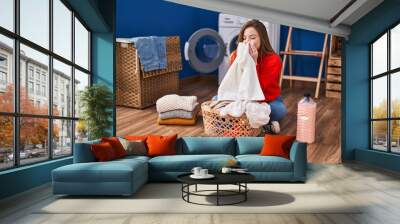 Young blonde woman smelling clothes sitting on floor at laundry room Wall mural