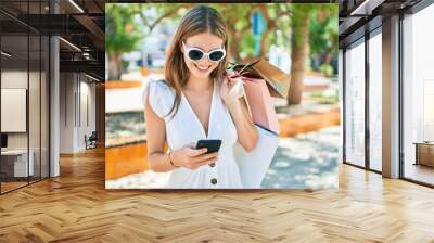 Young blonde woman smiling happy holding shopping bags and using smartphone at street of city Wall mural