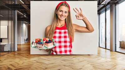 Young blonde woman wearing baker uniform holding homemade cakes doing ok sign with fingers, smiling friendly gesturing excellent symbol Wall mural