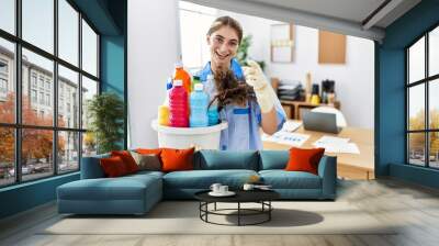 Young blonde woman wearing cleaner uniform holding cleaning products pointing to you and the camera with fingers, smiling positive and cheerful Wall mural