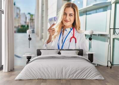 Young blonde woman wearing doctor uniform holding thermometer at hospital Wall mural