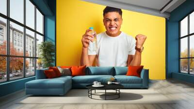 Young brazilian man holding bottle of water standing over isolated yellow background screaming proud and celebrating victory and success very excited, cheering emotion Wall mural