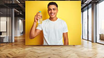 Young brazilian man holding bottle of water standing over isolated yellow background with a happy face standing and smiling with a confident smile showing teeth Wall mural