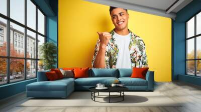 Young brazilian man on vacation wearing summer floral shirt over isolated yellow background smiling with happy face looking and pointing to the side with thumb up. Wall mural