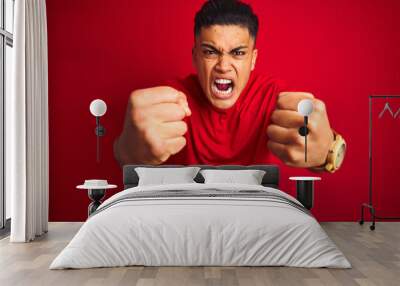 Young brazilian man wearing t-shirt standing over isolated red background angry and mad raising fists frustrated and furious while shouting with anger. Rage and aggressive concept. Wall mural