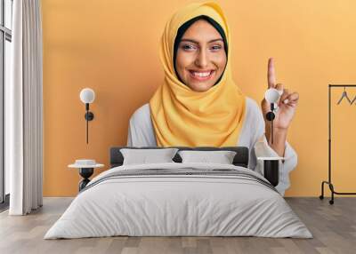 Young brunette arab woman wearing traditional islamic hijab scarf showing and pointing up with finger number one while smiling confident and happy. Wall mural