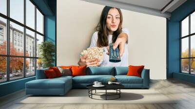 Young brunette woman eating popcorn using tv control looking at the camera blowing a kiss being lovely and sexy. love expression. Wall mural