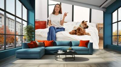 Young brunette woman sitting on the bed with two dogs smiling happy pointing with hand and finger to the side Wall mural