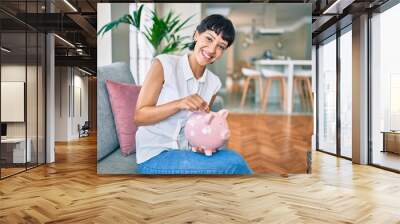 Young brunette woman smiling happy putting money savings inside of piggy bank Wall mural