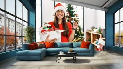 Young brunette woman standing by christmas tree holding decoration cheerful with a smile on face pointing with hand and finger up to the side with happy and natural expression Wall mural