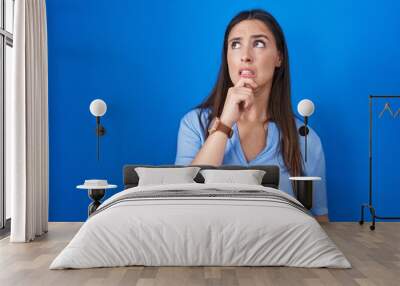 Young brunette woman standing over blue background thinking worried about a question, concerned and nervous with hand on chin Wall mural