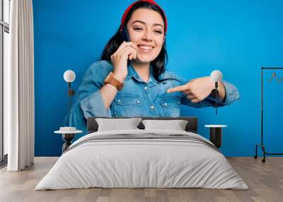 Young brunette woman talking on smartphone over blue isolated background with surprise face pointing finger to himself Wall mural