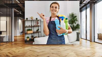 Young brunette woman wearing apron holding cleaning products smiling happy pointing with hand and finger to the side Wall mural