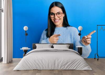 Young brunette woman wearing casual clothes and glasses smiling and looking at the camera pointing with two hands and fingers to the side. Wall mural