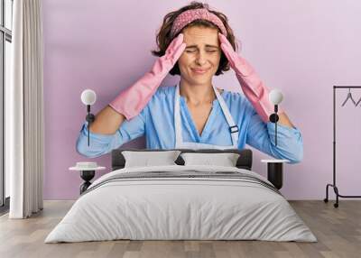 Young brunette woman wearing cleaner apron and gloves suffering from headache desperate and stressed because pain and migraine. hands on head. Wall mural