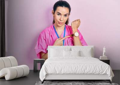 Young brunette woman wearing doctor uniform and stethoscope in hurry pointing to watch time, impatience, looking at the camera with relaxed expression Wall mural