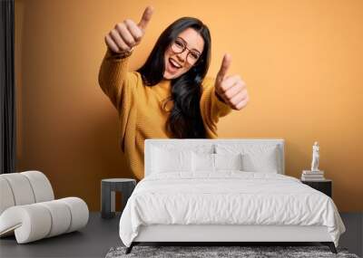 Young brunette woman wearing glasses and casual sweater over yellow isolated background approving doing positive gesture with hand, thumbs up smiling and happy for success. Winner gesture. Wall mural