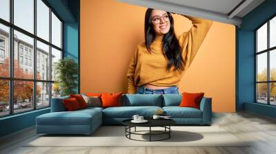 Young brunette woman wearing glasses and casual sweater over yellow isolated background smiling confident touching hair with hand up gesture, posing attractive and fashionable Wall mural