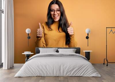 Young brunette woman wearing glasses and casual sweater over yellow isolated background success sign doing positive gesture with hand, thumbs up smiling and happy. Cheerful expression and winner Wall mural