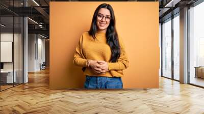 Young brunette woman wearing glasses and casual sweater over yellow isolated background with hands together and crossed fingers smiling relaxed and cheerful. Success and optimistic Wall mural