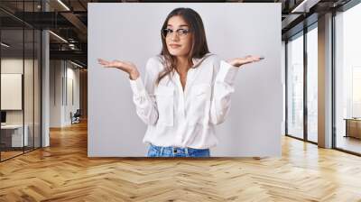 Young brunette woman wearing glasses clueless and confused expression with arms and hands raised. doubt concept. Wall mural