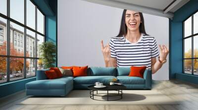 Young brunette woman wearing striped t shirt shouting with crazy expression doing rock symbol with hands up. music star. heavy music concept. Wall mural