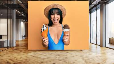 Young brunette woman with bangs wearing swimwear holding two ice cream cones celebrating crazy and amazed for success with open eyes screaming excited. Wall mural