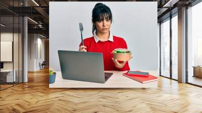 Young brunette woman with bangs working at the office eating healthy salad skeptic and nervous, frowning upset because of problem. negative person. Wall mural