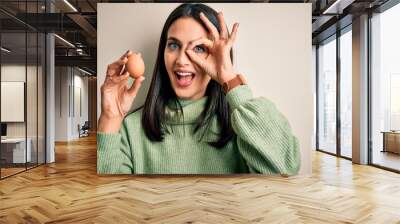 Young brunette woman with blue eyes holding fresh raw egg over isolated background with happy face smiling doing ok sign with hand on eye looking through fingers Wall mural