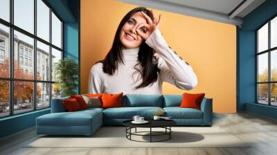 Young brunette woman with blue eyes wearing casual sweater over isolated yellow background doing ok gesture with hand smiling, eye looking through fingers with happy face. Wall mural