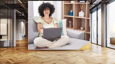 Young brunette woman with curly hair using laptop sitting on the sofa at home happy face smiling with crossed arms looking at the camera. positive person. Wall mural