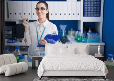 Young brunette woman working at scientist laboratory with a big smile on face, pointing with hand finger to the side looking at the camera. Wall mural