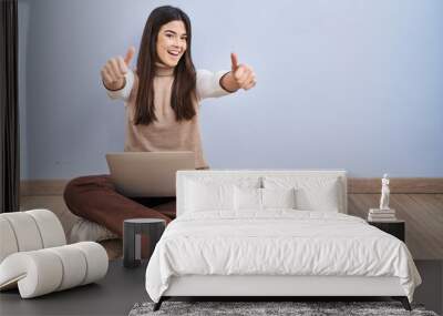 Young brunette woman working using computer laptop sitting on the floor approving doing positive gesture with hand, thumbs up smiling and happy for success. winner gesture. Wall mural