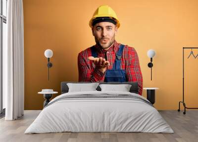 Young builder man wearing construction uniform and safety helmet over yellow isolated background looking at the camera blowing a kiss with hand on air being lovely and sexy. Love expression. Wall mural