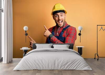 Young builder man wearing construction uniform and safety helmet over yellow isolated background smiling and looking at the camera pointing with two hands and fingers to the side. Wall mural