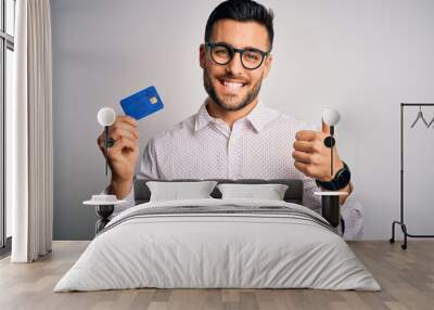 Young business man holding credit card over isolated background happy with big smile doing ok sign, thumb up with fingers, excellent sign Wall mural