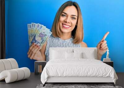 Young caucasian blonde woman holding 100 romanian leu banknotes smiling happy pointing with hand and finger to the side Wall mural