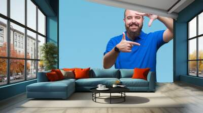 Young caucasian hipster man wearing blue shirt over isolated background smiling making frame with hands and fingers with happy face. Creativity and photography concept. Wall mural