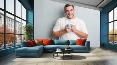 Young caucasian man over grey grunge wall driking a cup of coffee with a happy face standing and smiling with a confident smile showing teeth Wall mural