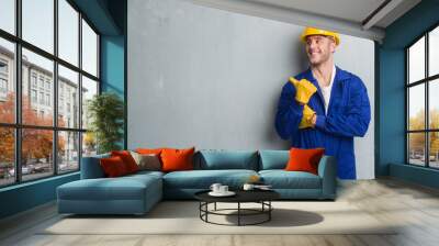 Young caucasian man over grey grunge wall wearing contractor uniform and safety helmet pointing and showing with thumb up to the side with happy face smiling Wall mural