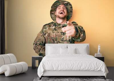 Young caucasian man wearing camouflage army uniform laughing at you, pointing finger to the camera with hand over body, shame expression Wall mural