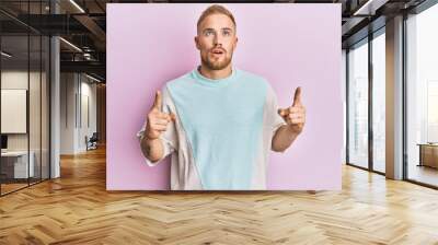 Young caucasian man wearing casual clothes amazed and surprised looking up and pointing with fingers and raised arms. Wall mural