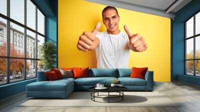 Young caucasian man wearing casual white t-shirt over yellow isolated background approving doing positive gesture with hand, thumbs up smiling and happy for success. Winner gesture. Wall mural