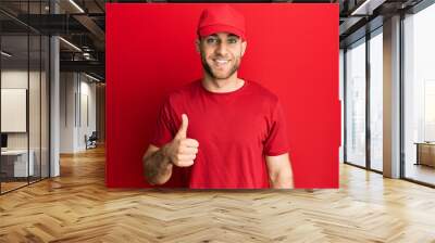 Young caucasian man wearing delivery uniform and cap smiling happy and positive, thumb up doing excellent and approval sign Wall mural