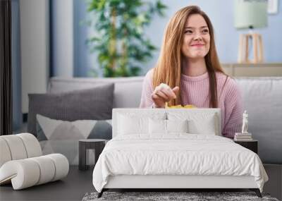 Young caucasian woman eating chips potatoes sitting on sofa at home Wall mural